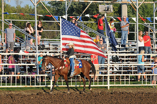 A rodeo queen holding a large American flag while riding a brown horse